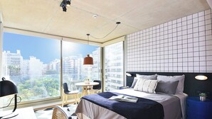 A modern bedroom with a large window, a bed with a blue headboard, a desk with a chair, and a view of the cityscape.