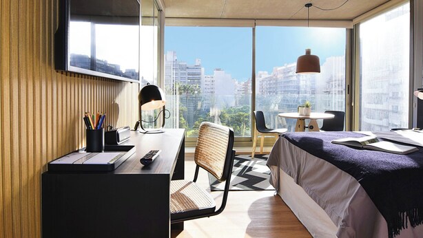 A modern hotel room with a large window, a desk with a lamp, a chair, a bed, and a small table with a plant.