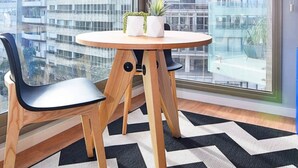A modern room with a round wooden table, two black chairs, a hanging orange pendant light, and a view of high-rise buildings through large windows.