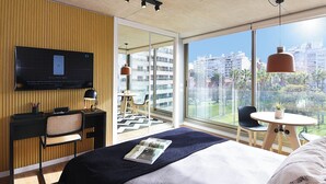 A modern hotel room with a large bed, a desk with a chair, a TV mounted on the wall, and a balcony with a view of the city.