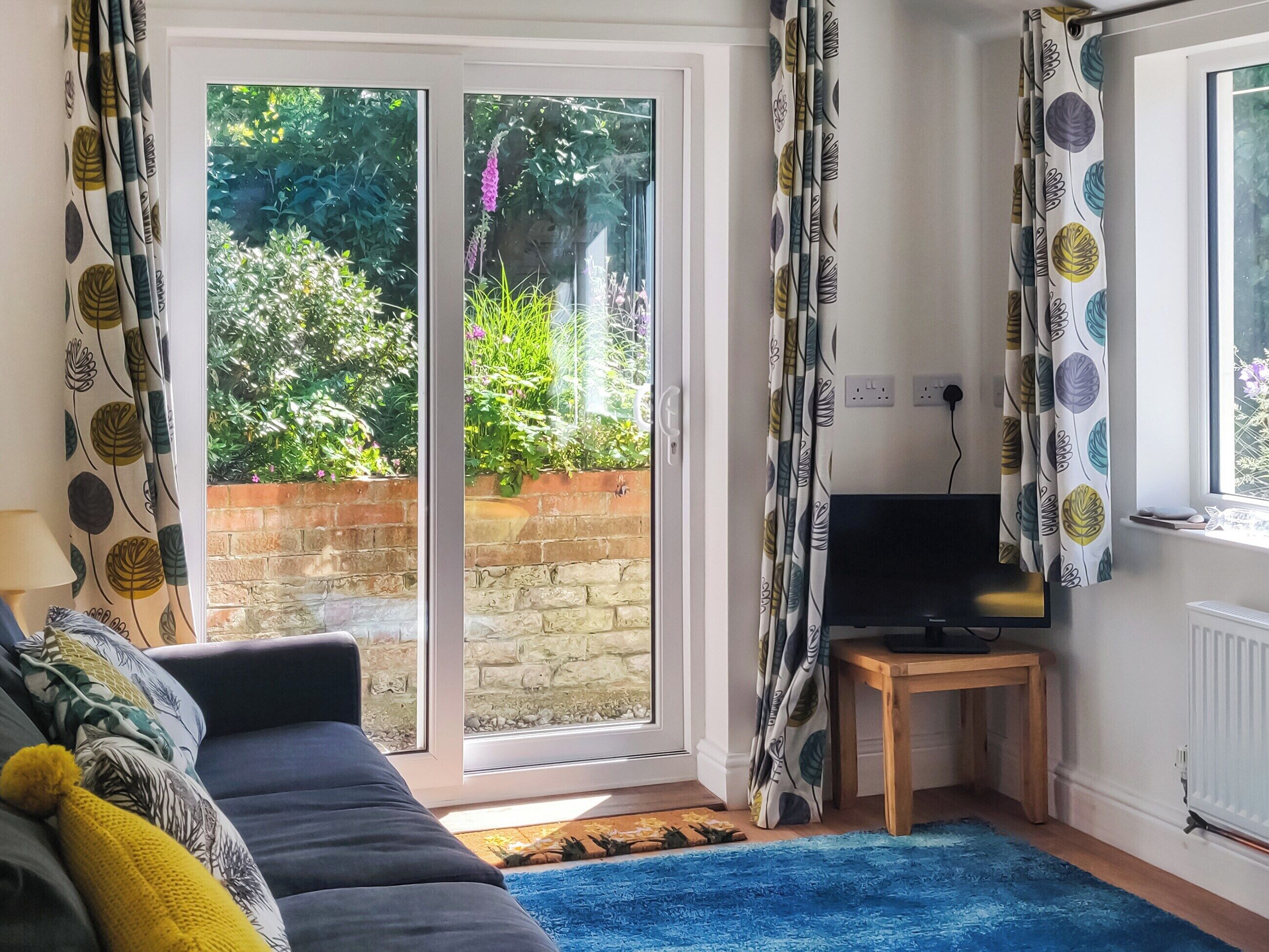 A living room with a sofa, a TV on a wooden stand, and a sliding glass door leading to a garden.