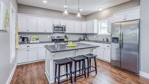 A modern kitchen with white cabinets, a large island, bar stools, a stainless steel refrigerator, and pendant lighting.