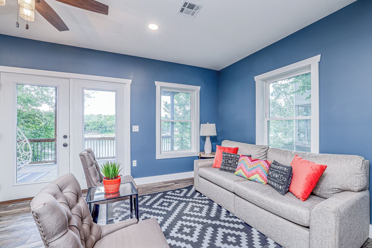 A living room with a grey sofa, a glass coffee table, and a patterned rug. There are two windows with white frames and a ceiling fan.