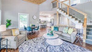 A spacious living room with a high ceiling, a staircase, and a kitchen in the background.