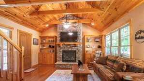 A cozy living room with a stone fireplace, wooden ceiling beams, a television, and a large window. - Redbud Retreat ~ Rustic family friendly cabin with New Hot Tub in Townsend! (Townsend)