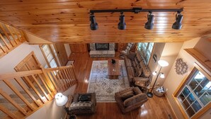 A spacious loft-style living area with a high ceiling, wooden beams, a stone fireplace, a large sofa, a coffee table, and a view of the outdoors through large windows. - Redbud Retreat ~ Rustic family friendly cabin with New Hot Tub in Townsend! (Townsend)