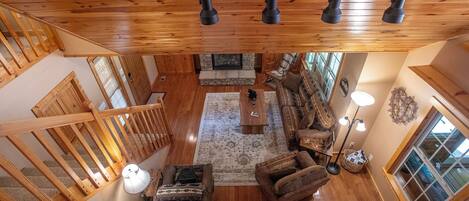 A spacious loft-style living area with a high ceiling, wooden beams, a stone fireplace, a large sofa, a coffee table, and a view of the outdoors through large windows.
