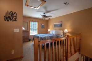 A bedroom with a bed, two bedside lamps, a ceiling fan, a window with blinds, a painting on the wall, and a 'Go Wild' sign. - Redbud Retreat ~ Rustic family friendly cabin with New Hot Tub in Townsend! (Townsend)