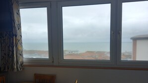Une pièce avec une table ronde en bois et quatre chaises, une fenêtre donnant sur la mer et les bâtiments, et un rideau sur le côté gauche.
