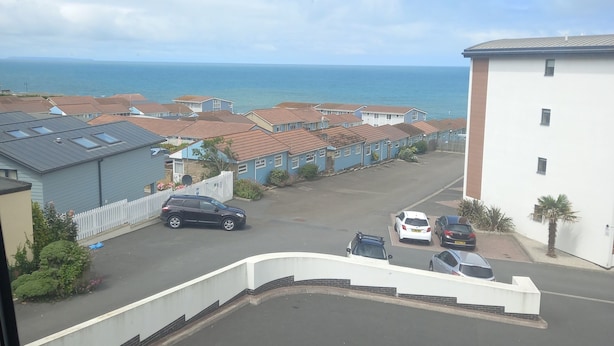 Un quartier résidentiel offrant une vue sur la mer, avec des maisons aux toits rouges, un bâtiment blanc et des voitures garées.