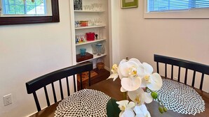 Un coin repas comprenant une table en bois, deux chaises, un lustre, un miroir, une fenêtre avec des stores et une étagère où sont rangés des verres.