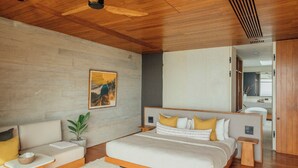 Quarto moderno com teto de madeira, cama grande com roupa de cama branca e almofadas amarelas, sofá, mesa de apoio e planta em vaso.