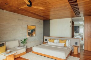 A modern bedroom with a wooden ceiling, a large bed with multiple pillows, a sofa, a small table, and a potted plant.
