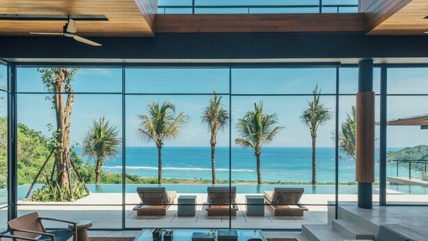 Uma sala de estar moderna com uma grande parede de vidro com vista para uma piscina e para o oceano, equipada com um sofá, cadeiras e uma mesa de centro.