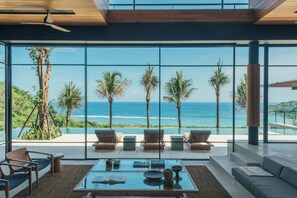 Un salon moderne doté d’une grande baie vitrée donnant sur une piscine et offrant une vue sur l’océan, et aménagé avec un canapé, des fauteuils et une table basse.