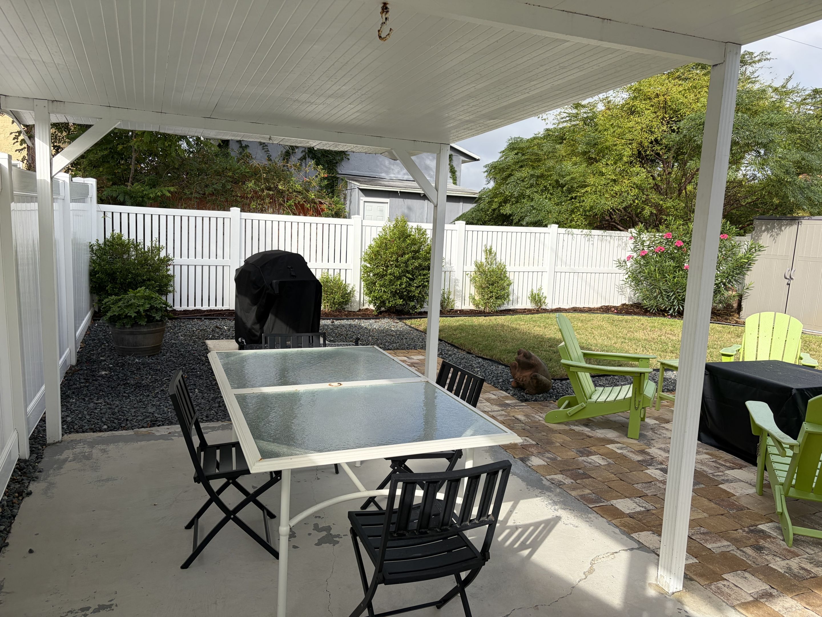 A covered patio area with a glass-top table, black chairs, a grill, and a white fence in the background.