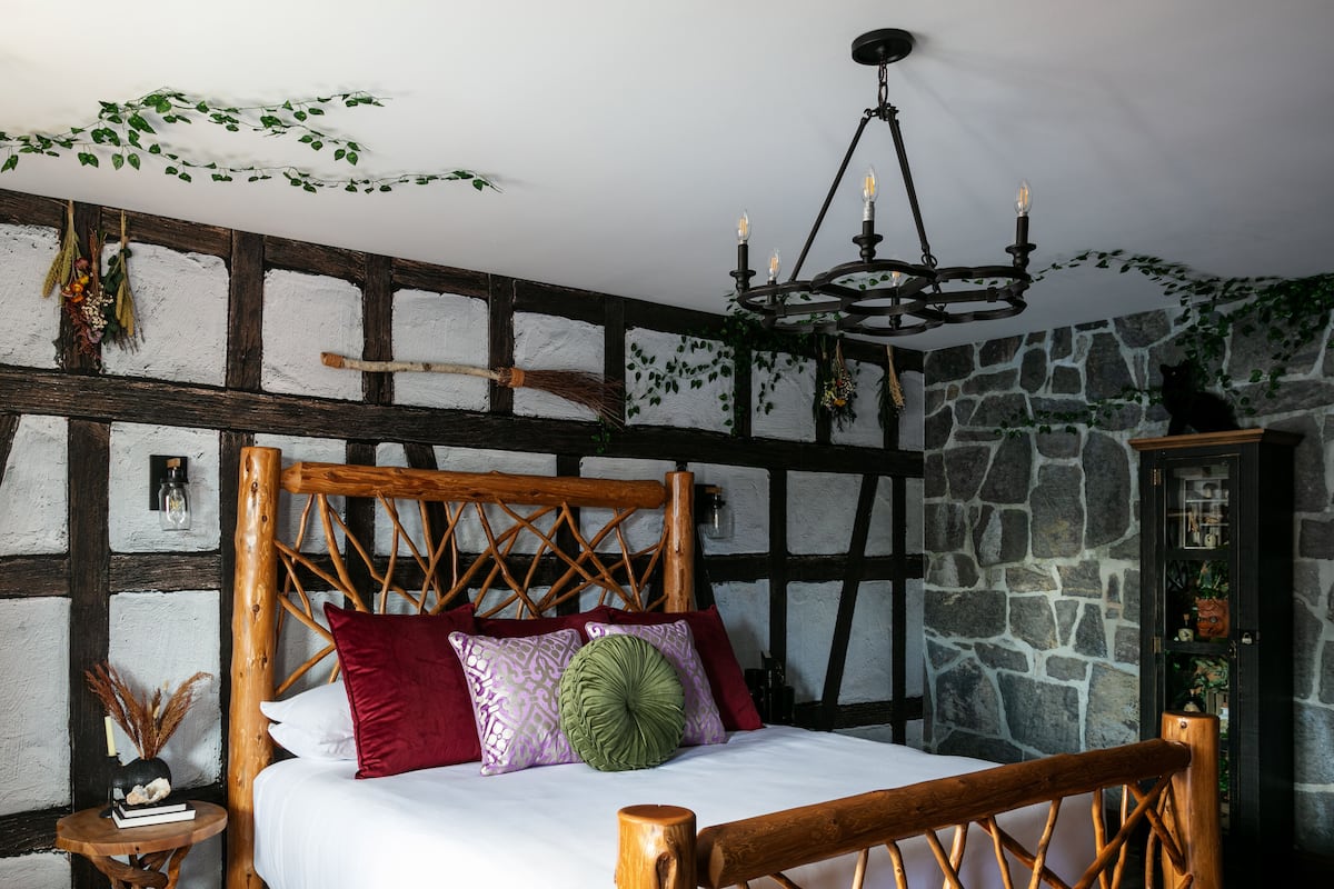 A wooden four-poster bed with white linens, a wooden nightstand with a book and a vase, and a stone wall with a stone cabinet.