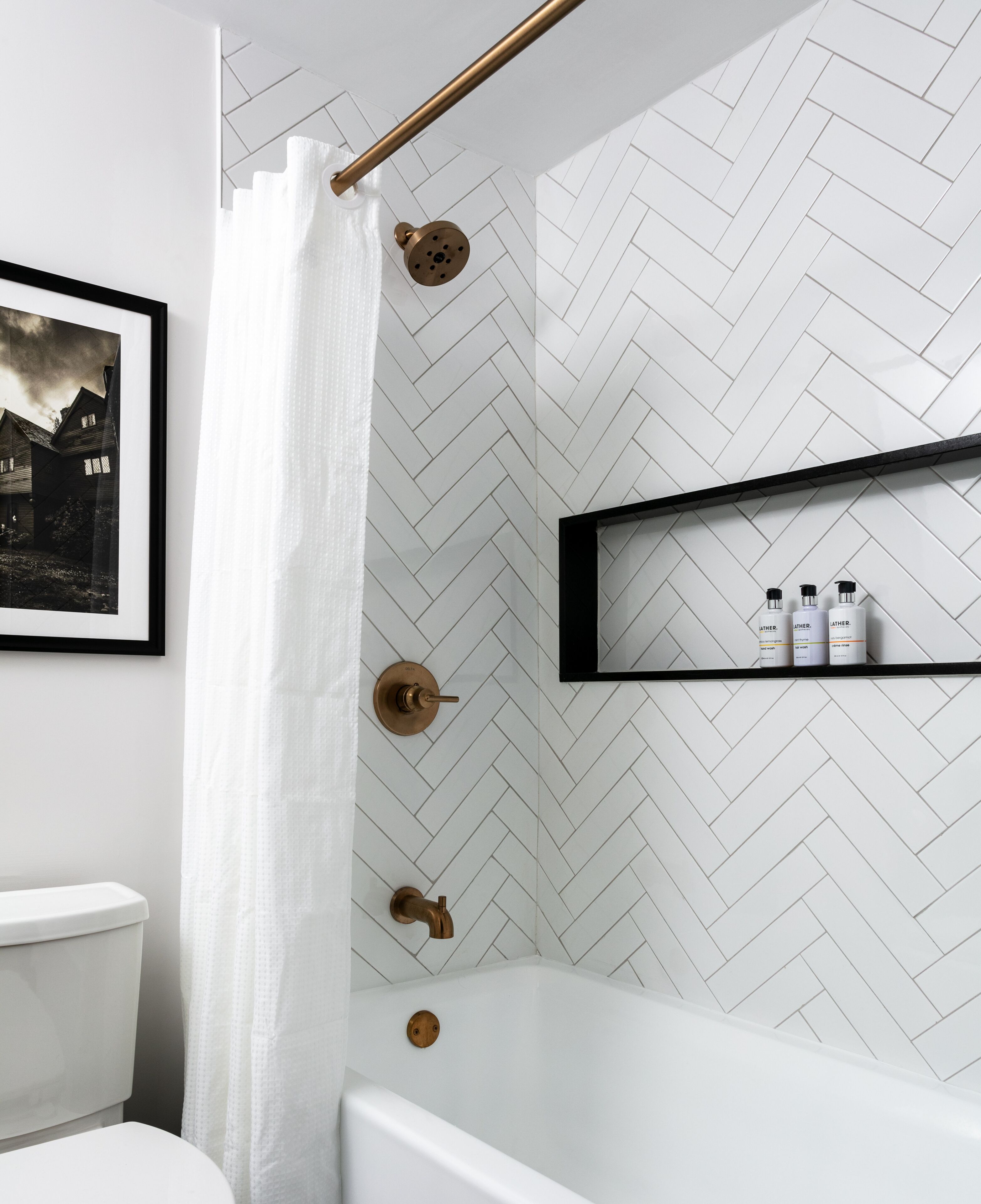 A modern bathroom with a herringbone tile pattern, a white bathtub, a shower with a gold showerhead, and a shelf with toiletries.
