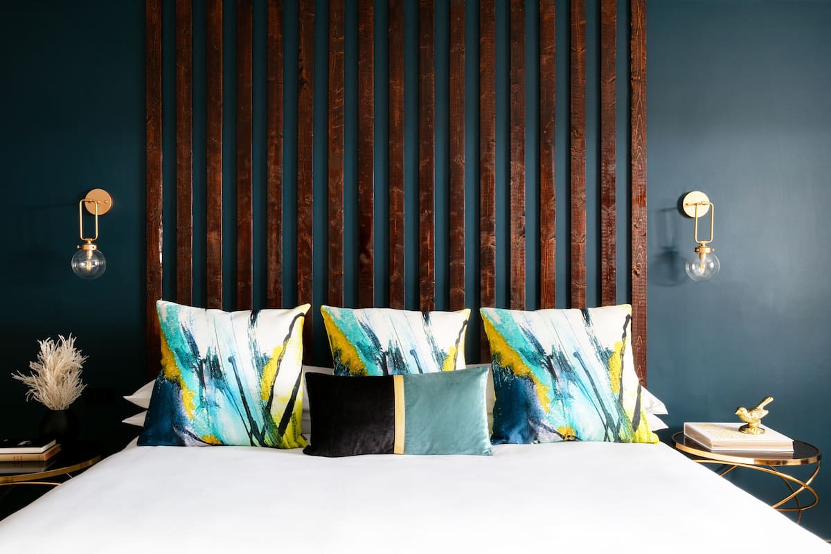 A bed with colorful pillows, a dark wooden headboard, and two wall-mounted lamps with exposed bulbs.