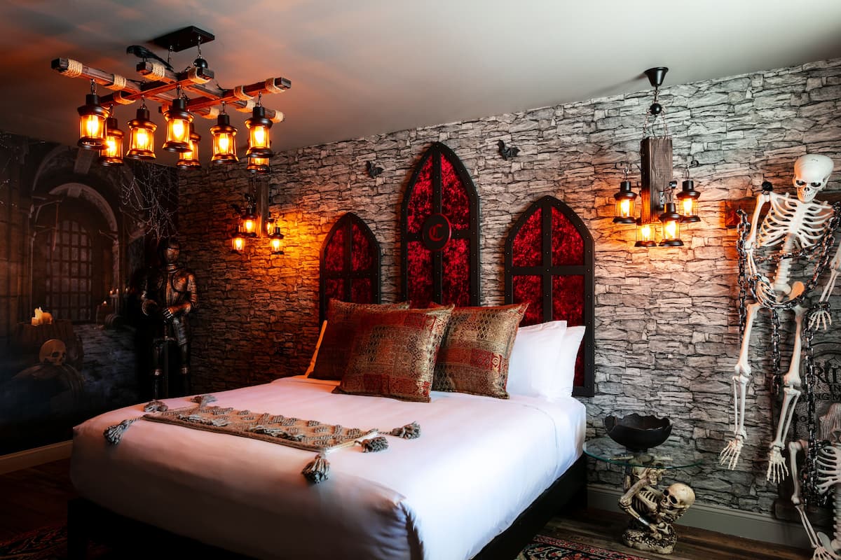 A bedroom with a stone wall, a bed with patterned pillows, a skeleton decoration, and a chandelier.