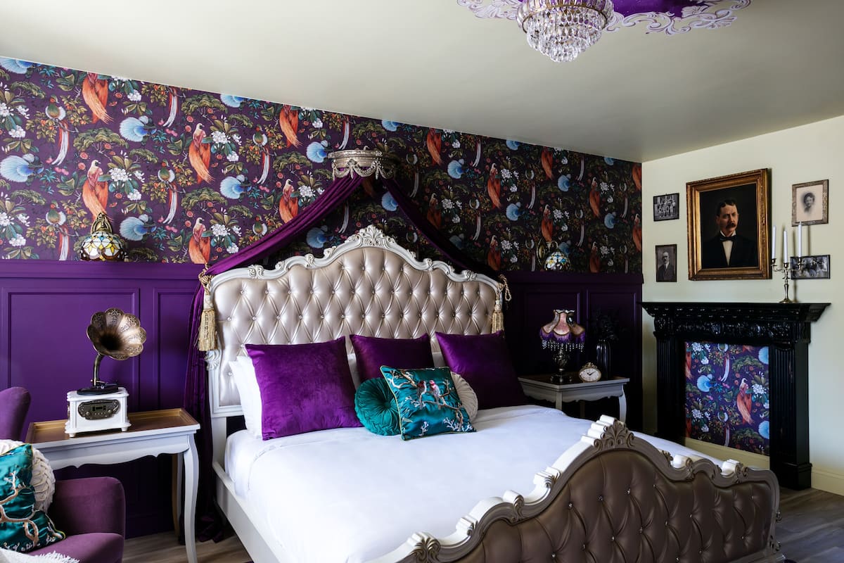 A bedroom with a tufted headboard, a bed with white and purple bedding, a vintage gramophone, a purple armchair, and a fireplace with a floral tapestry.