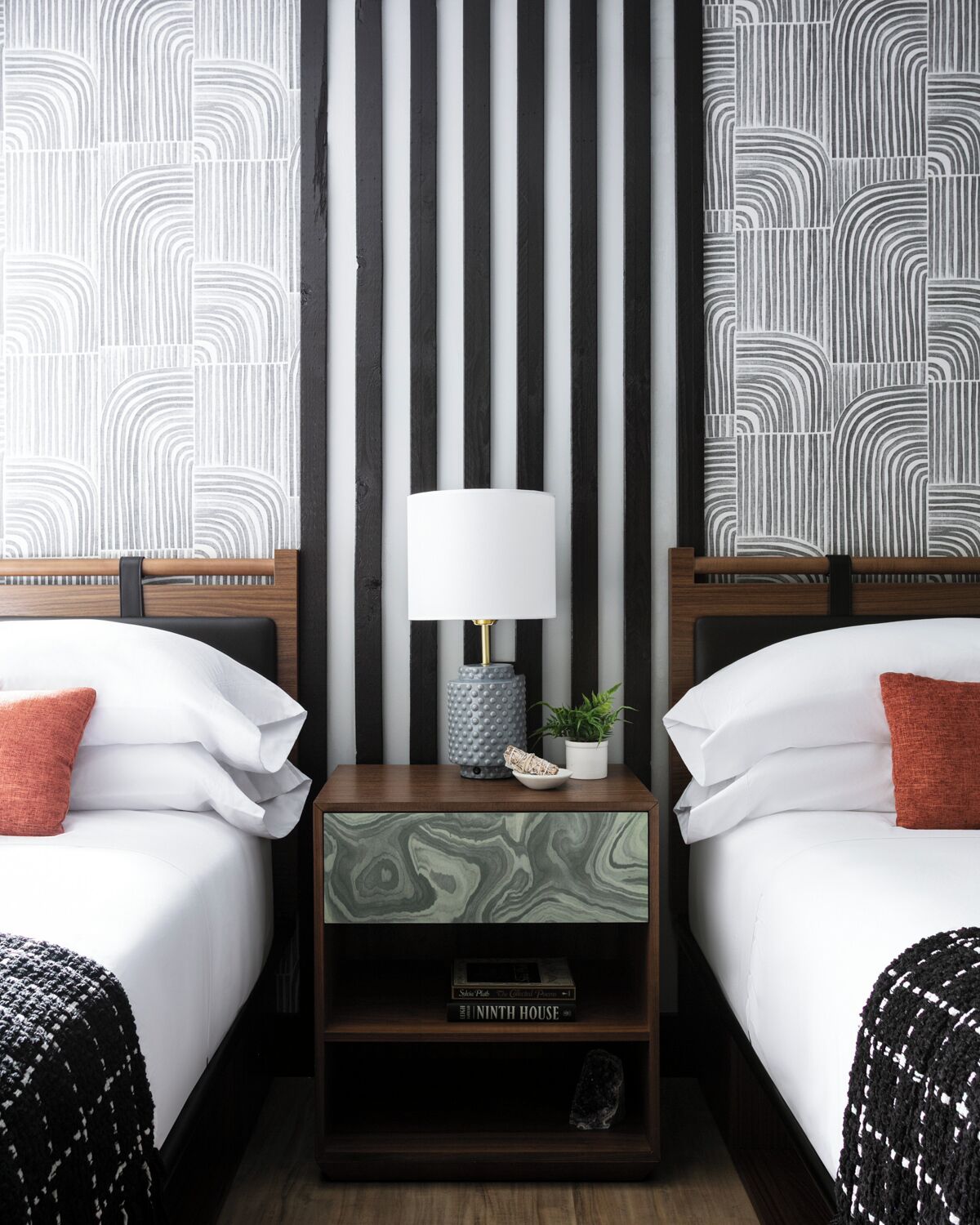 A hotel room with two beds, a nightstand with a lamp, a small plant, and a bookshelf with a book titled "The Ninth House".