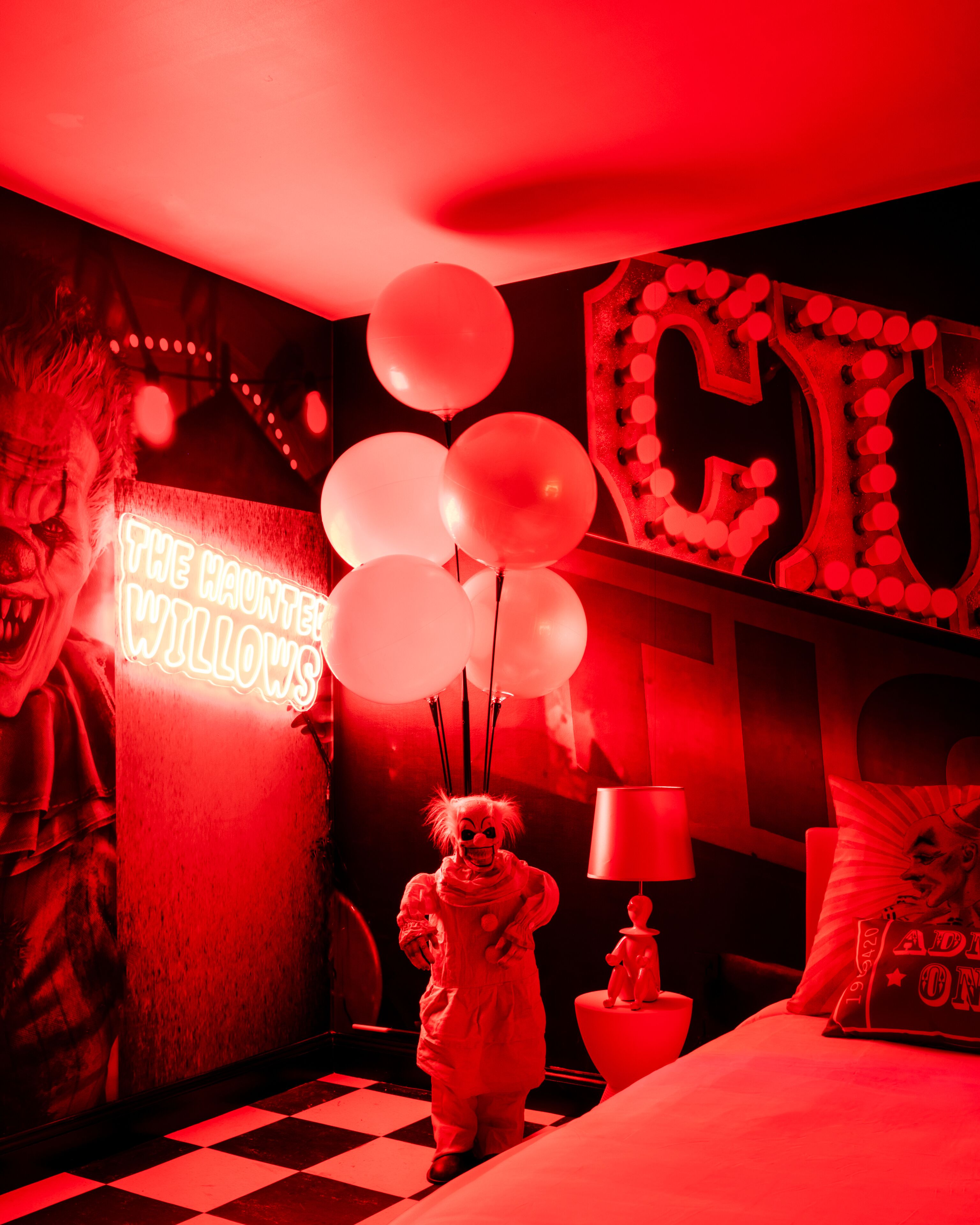 Una habitación con piso a cuadros, un letrero de neón que dice 'THE HAUNTED WILLOWS' y una figura con disfraz de payaso sosteniendo globos.