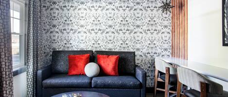 Una sala de estar moderna con un sofá oscuro, cojines rojos, una mesa de centro redonda y un comedor con una mesa blanca y sillas de madera.