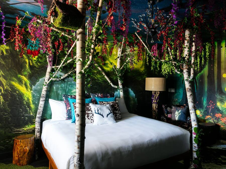 Een bed met witte beddengoed, omgeven door berkenbomen en hangende planten, in een kamer met een muurschildering van een bos.