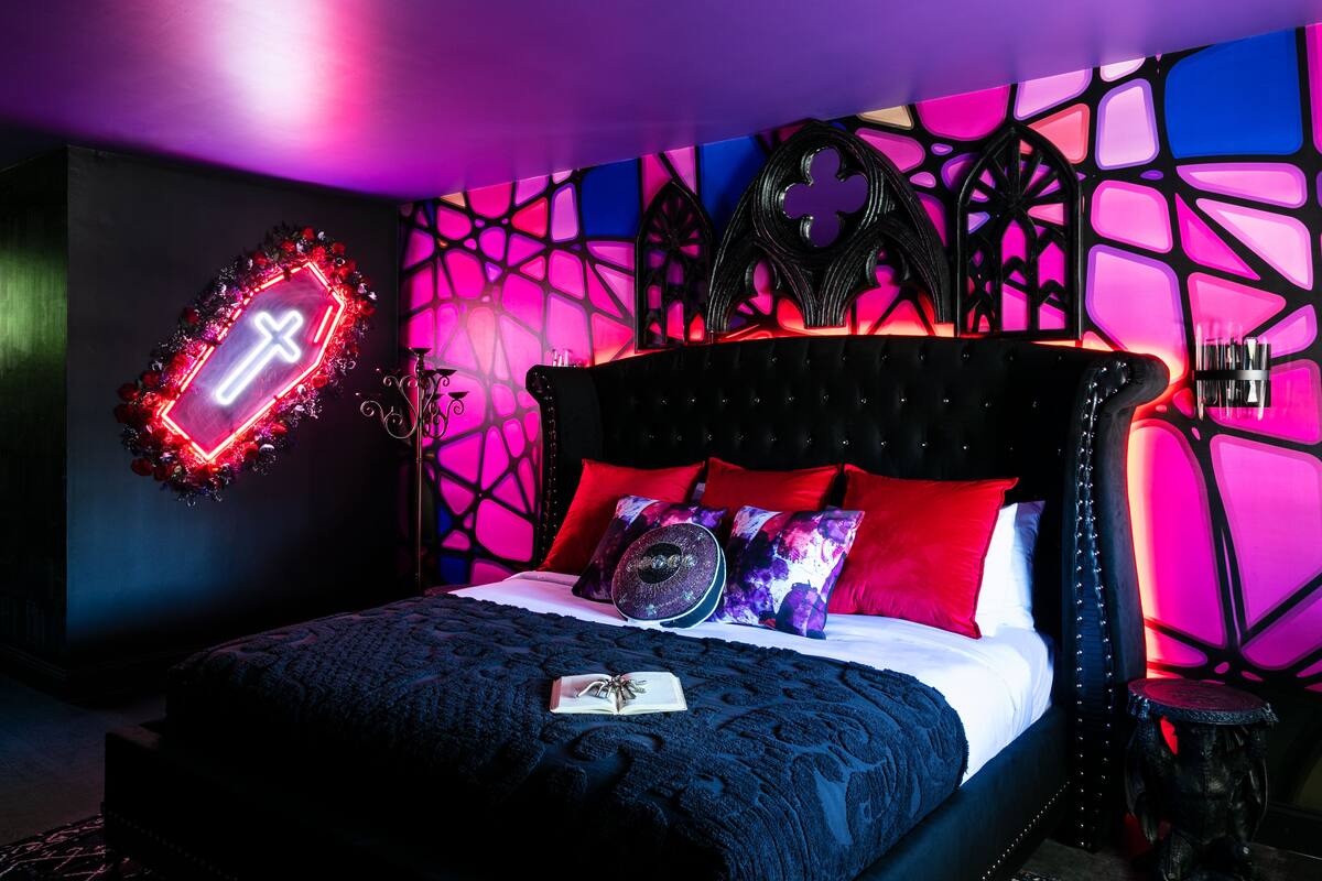A bedroom with a bed, a neon cross sign, and a geometric wall pattern.