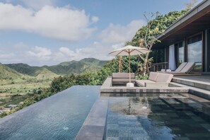 An infinity pool with a view of a mountainous landscape, a lounge area with a canopy, and a modern building structure.