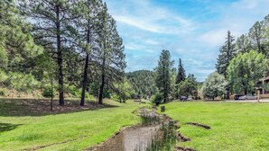 A grassy area with a small stream, surrounded by trees and a building in the background. - Hunter's Lodge by Ruidoso Vacation Rentals (Ruidoso)