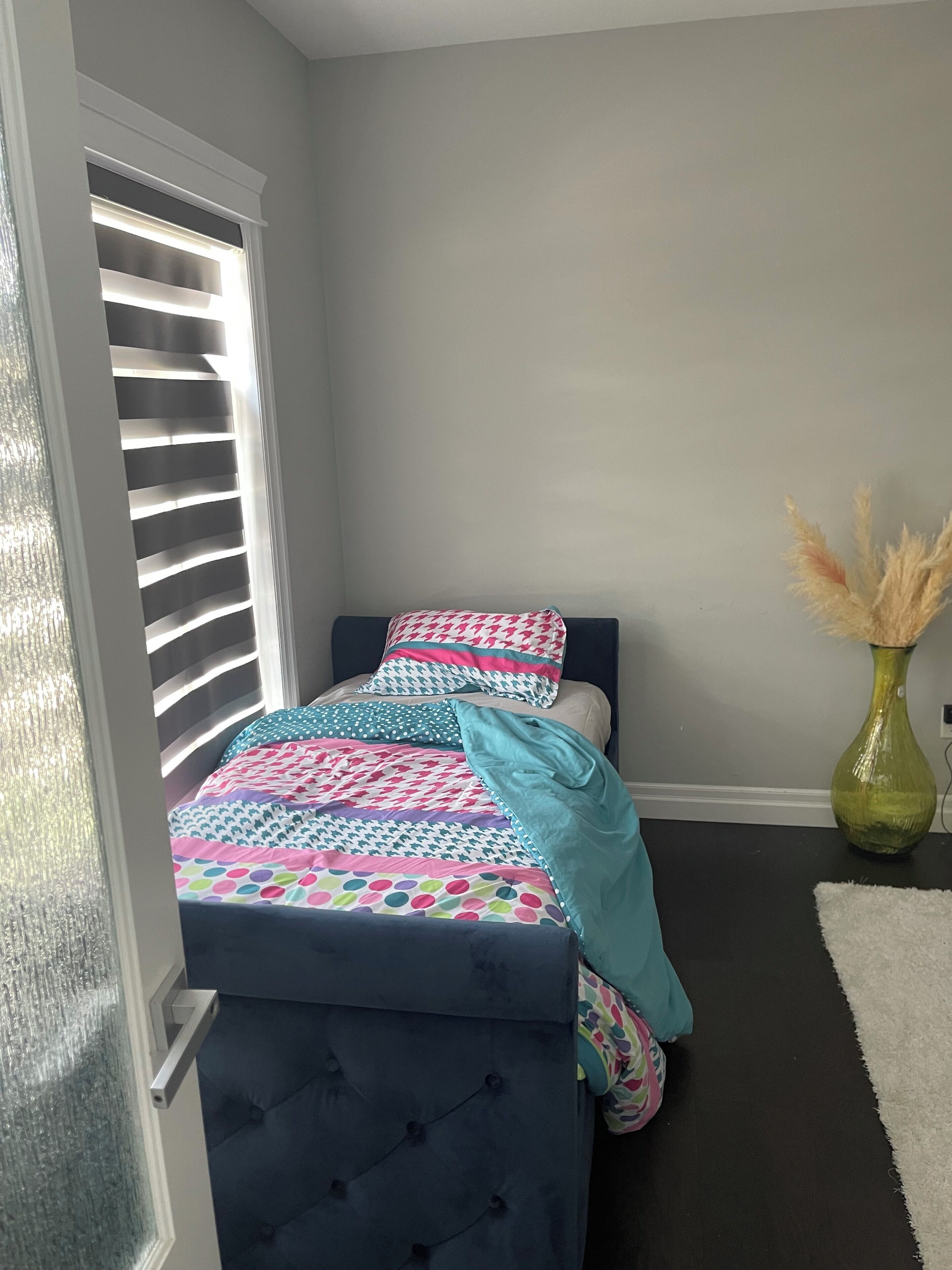 A small, neatly arranged bedroom with a bed, a window with blinds, a vase with dried flowers, and a white rug.