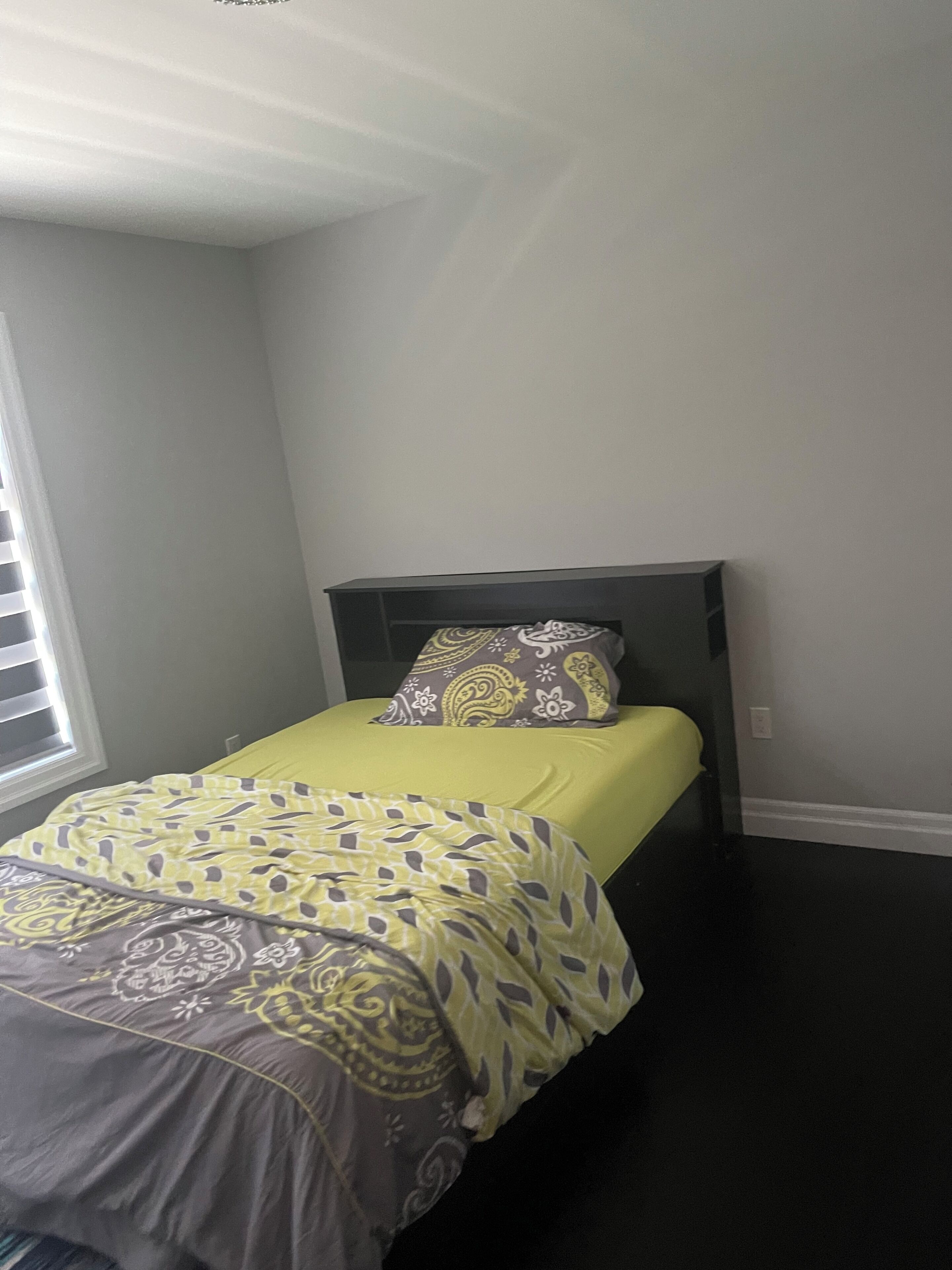 Un lit simple avec une couvre-lit à motifs et des oreillers, une tête de lit sombre et une fenêtre avec des stores.