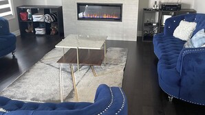 A modern living room with a fireplace, blue sofas, a coffee table, and shelves with decorative items. - Luxury getaway with pool and hot tub - family retreat  (Windsor)