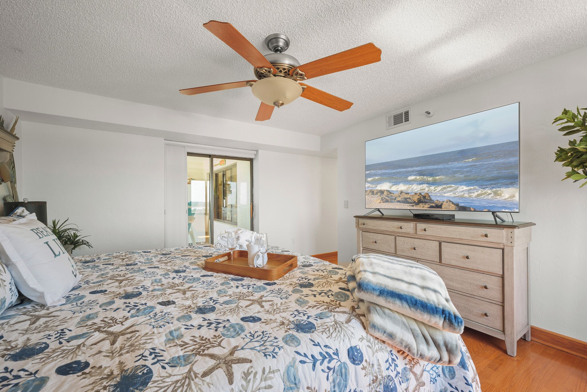 A bedroom with a bed, a ceiling fan, a dresser, a television, and a view of the ocean.