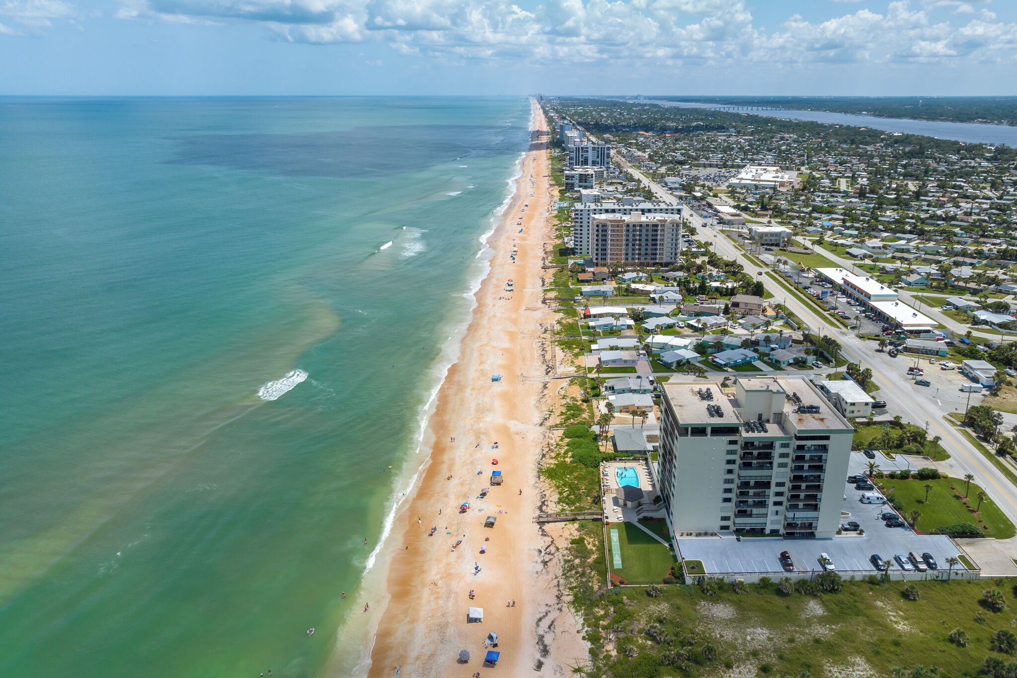 An aerial view of a coastal area with a beach, buildings, and a road.