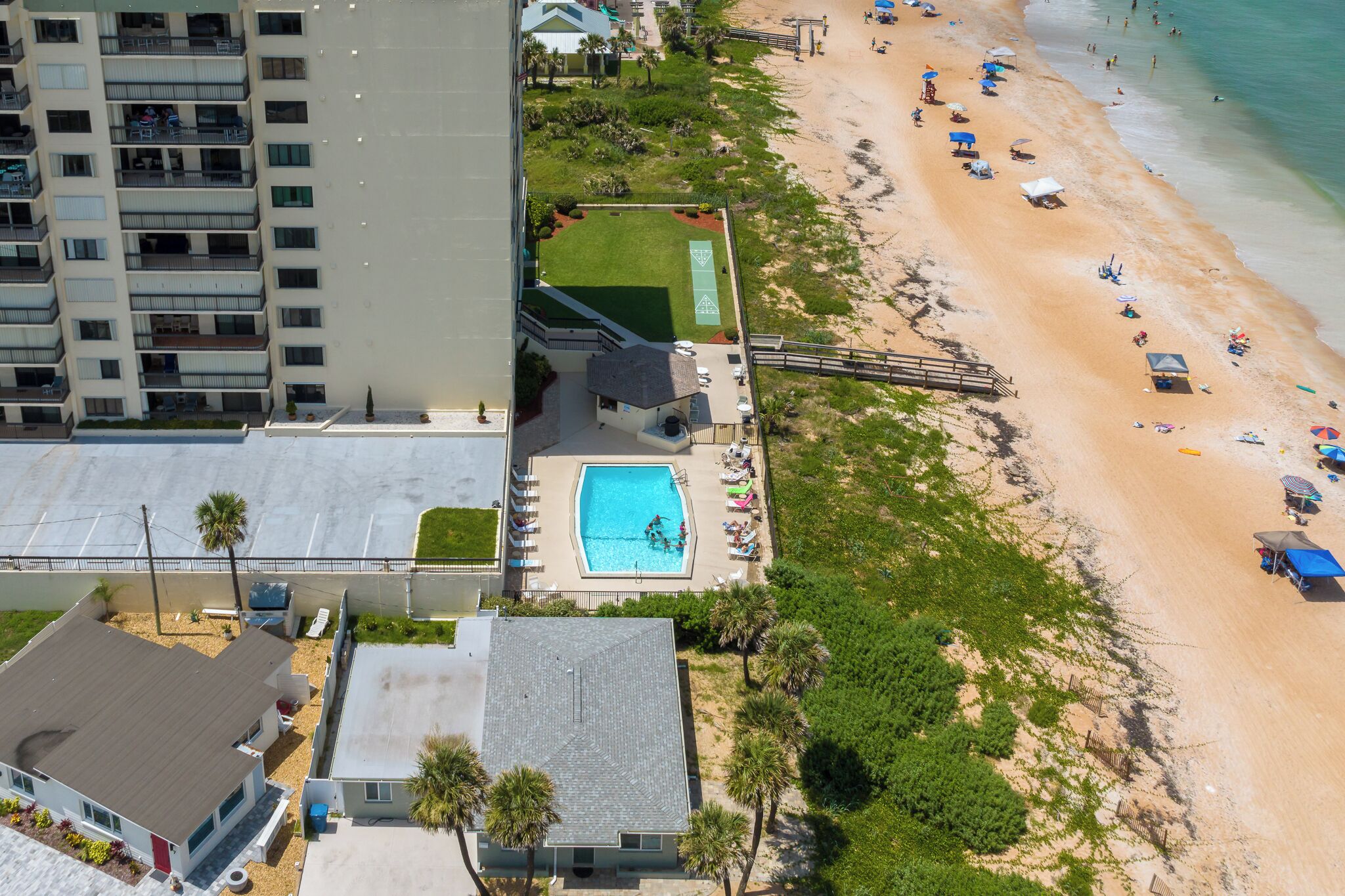 Vista aérea de una propiedad frente a la playa con piscina, sombrillas y un edificio de varios pisos al fondo.