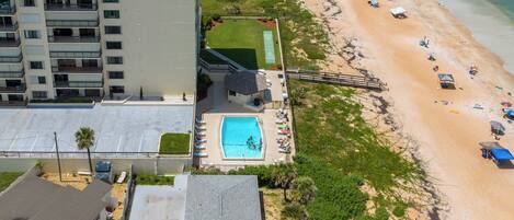 Vista aérea de una propiedad frente a la playa con piscina, sombrillas y un edificio de varios pisos al fondo.