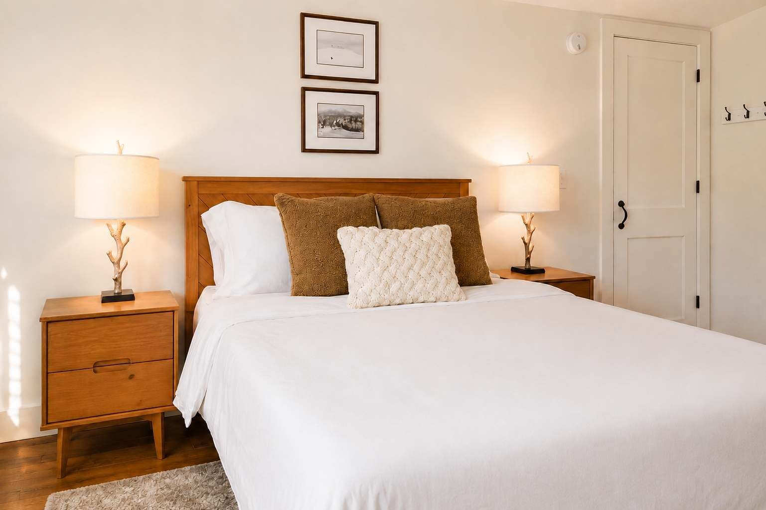 A neatly made bed with a wooden headboard, two bedside tables with lamps, a white door, and a wall-mounted picture.
