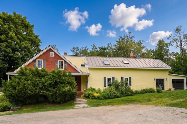 Une maison à deux étages, dont une partie est en briques rouges et l’autre est recouverte de panneaux jaunes, avec un porche à l’avant et une allée menant au garage.