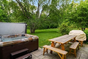 Un bain à remous extérieur, agrémenté d’une table et de bancs en bois, entouré d’une pelouse verdoyante et d’arbres.