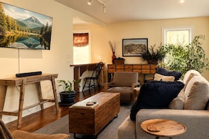 A living room with a sofa, coffee table, and a TV mounted on the wall. There is a wooden desk with a plant on it, and a chair in the corner.