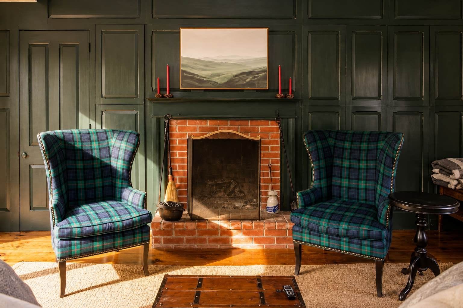 A room with a fireplace, two plaid armchairs, a small table, and a painting on the wall.