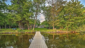 A wooden dock extending into a calm lake, surrounded by lush green trees and a house in the background.