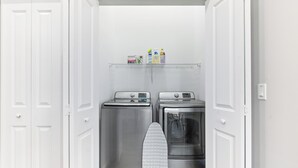 Un cuarto de lavado con dos lavadoras y secadoras de acero inoxidable, un estante con productos de limpieza y una plancha.