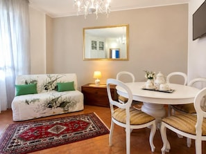 Un salon comprenant un canapé, une table à manger avec des chaises, un lustre, un miroir et un tapis à motifs.