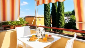 Un balcone con un tavolo apparecchiato per la colazione, con croissant e succo d'arancia, affacciato su una zona residenziale con alberi e palazzi.