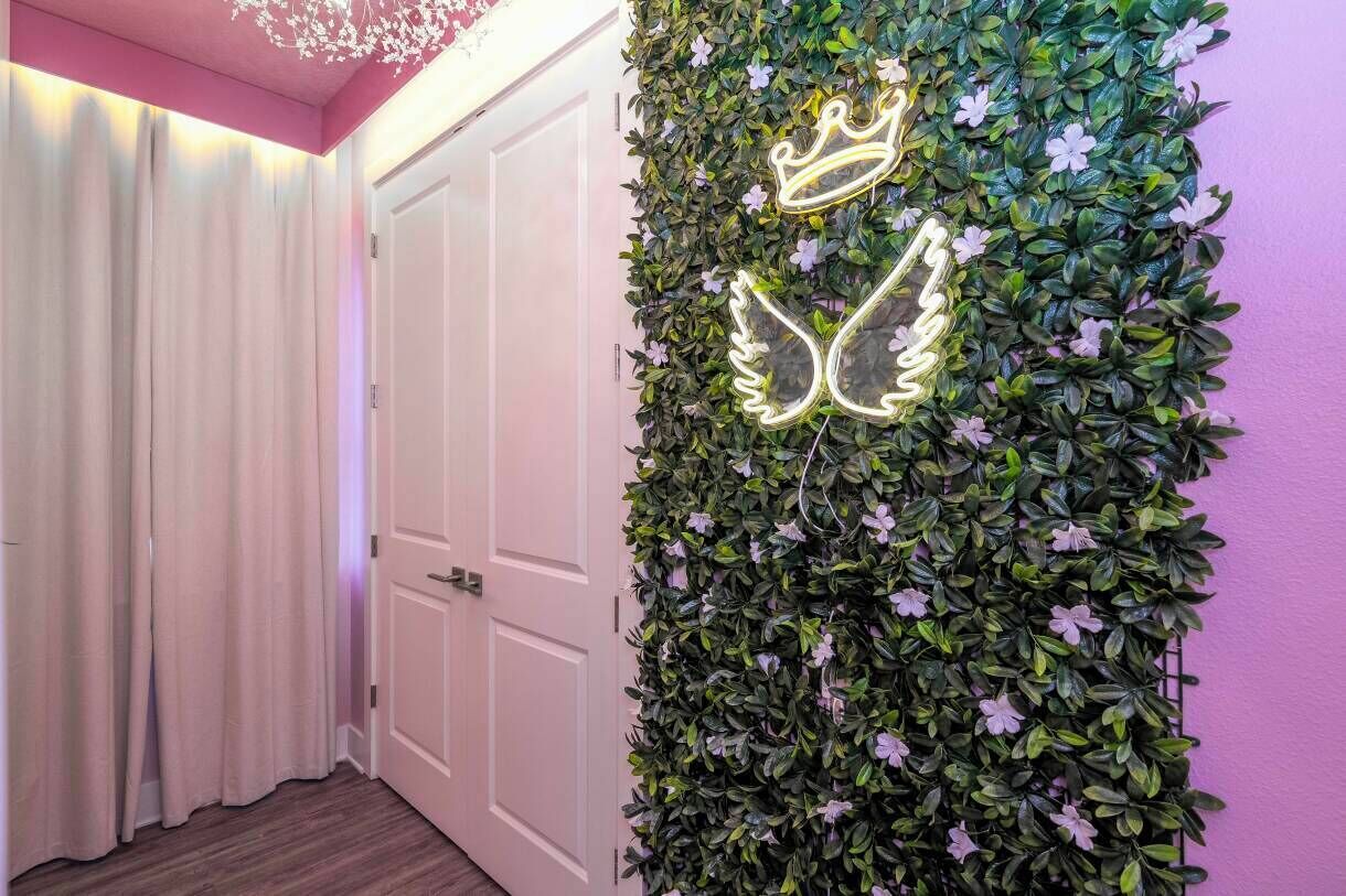 A room with a white door, a wall covered in green foliage and purple flowers, and a neon crown and wing design.