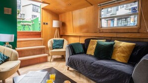 A cozy living room with a dark sofa, a coffee table with breakfast items, a lamp, and a view of the outside. - 4* Luxury floating wide beam houseboat moored in Edinburgh city center. (Edinburgh)